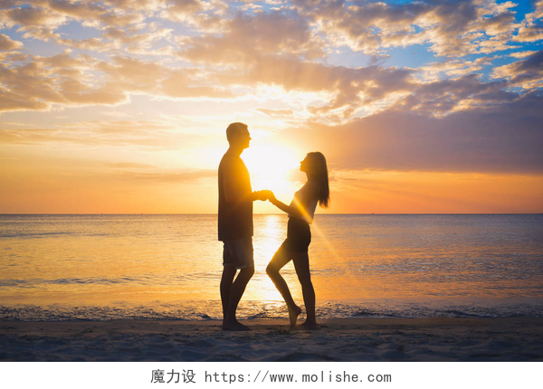 黄昏下站在沙滩手拉手的情侣在夕阳的暑假里，年轻漂亮的人在爱走，男人女人控股手海洋度假旅行夫妇在海滩上